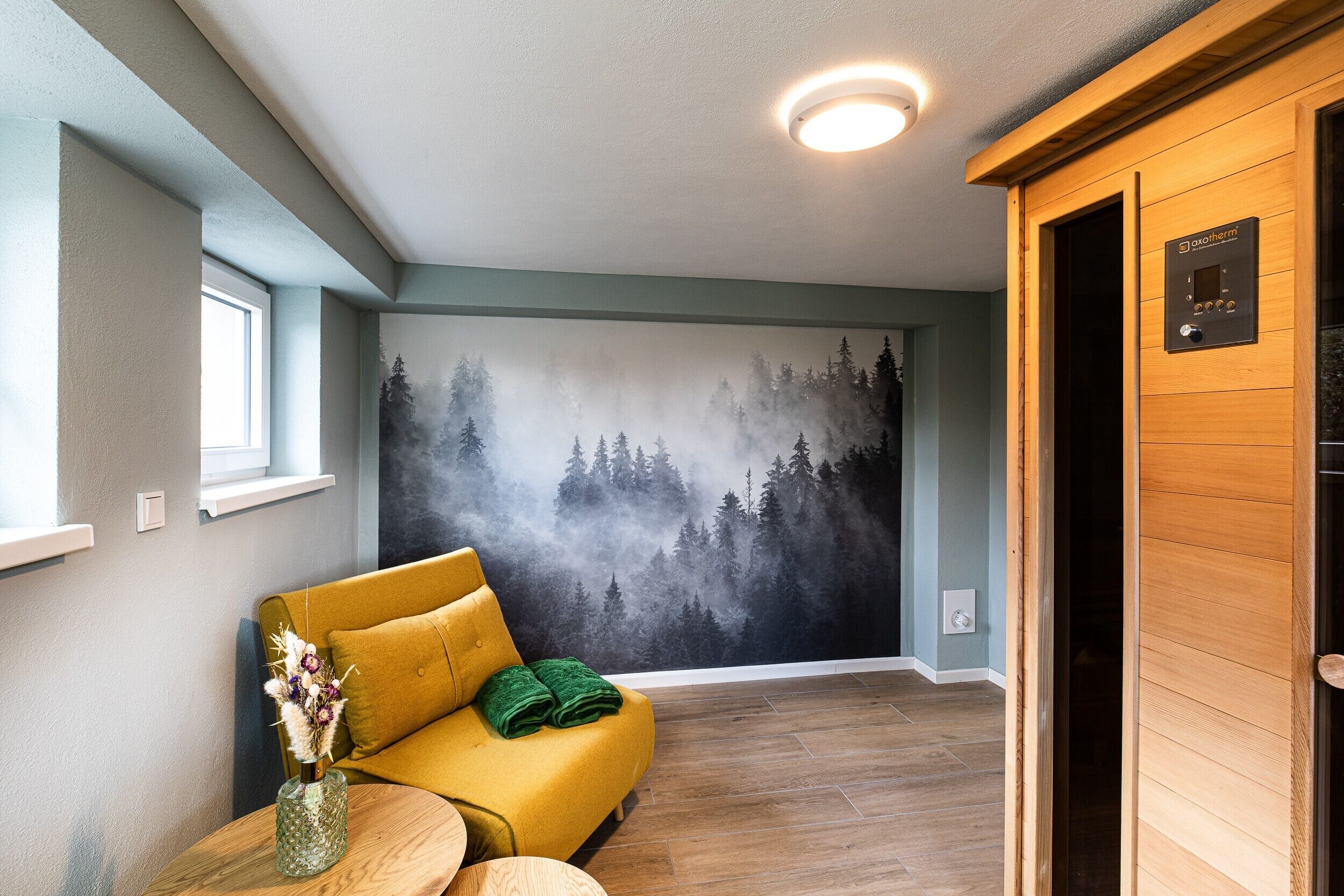 Ein Zimmer mit einem gelben Sofa, Holzboden, einem Wandgemälde eines Waldes und einer Deckenleuchte.