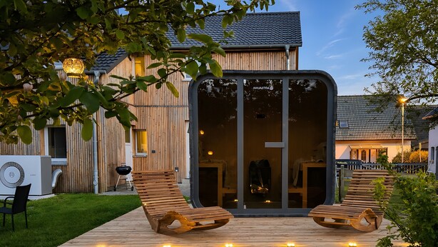 A modern house with a large glass door, a wooden deck with two lounge chairs, and a grill area.