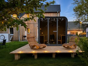 A modern house with a large glass door, a wooden deck with two lounge chairs, and a grill area. - Holiday Apartment 'Urlaub auf dem Land' with Private Terrace, Garden and Sauna Access (Feldberger Seenlandschaft)
