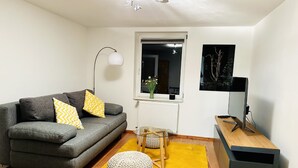 A modern living room with a grey sofa, yellow cushions, a round woven ottoman, a yellow rug, a wooden side table, a black TV stand, and a window with a view of flowers.