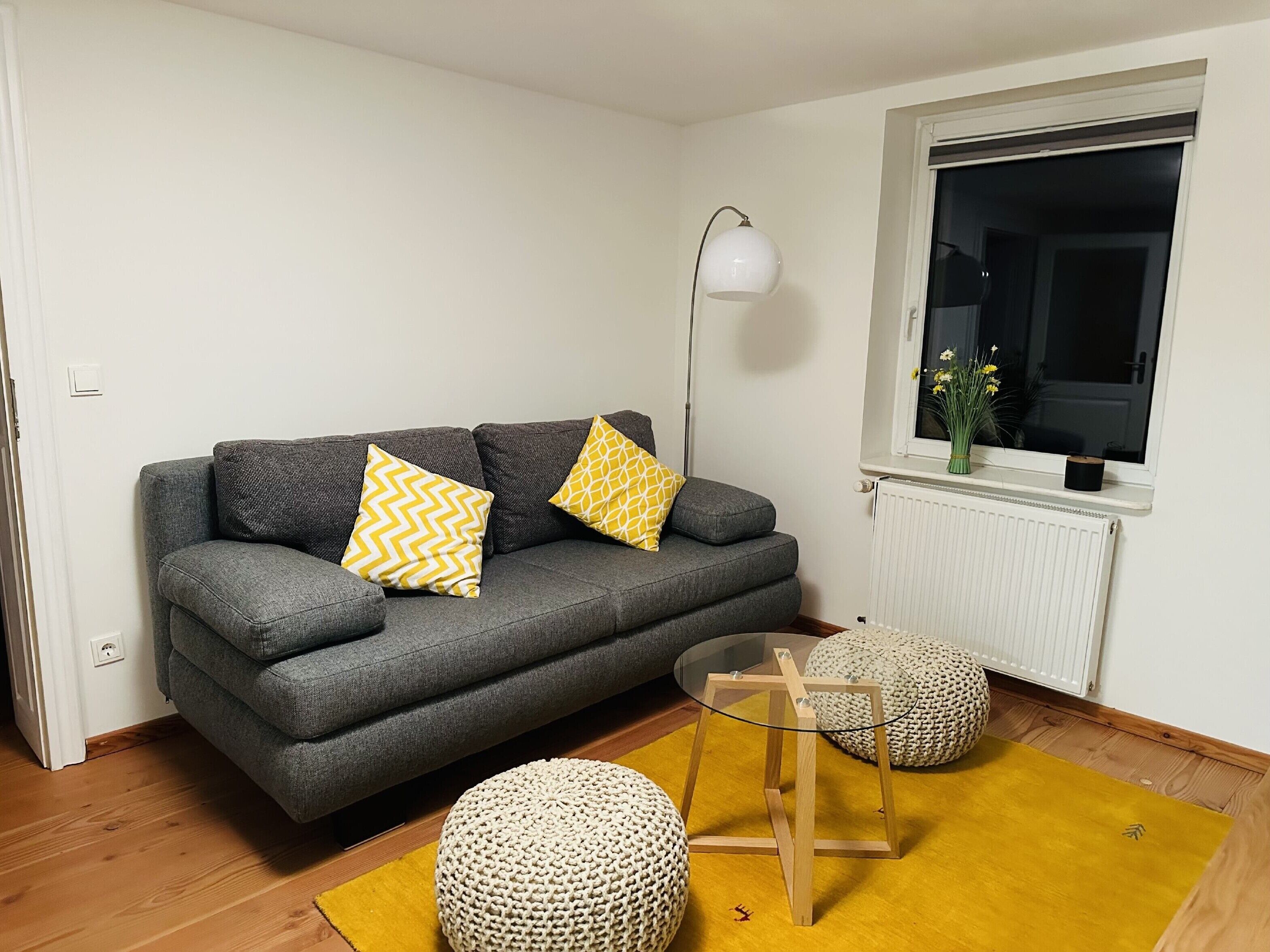 Ein graues Sofa mit gelb gemusterten Kissen, eine Stehlampe, ein runder gewebter Hocker und ein kleiner Glastisch.