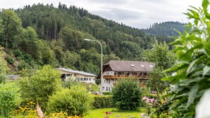 Paisagem rural com uma casa rodeada por vegetação exuberante e uma colina arborizada ao fundo.