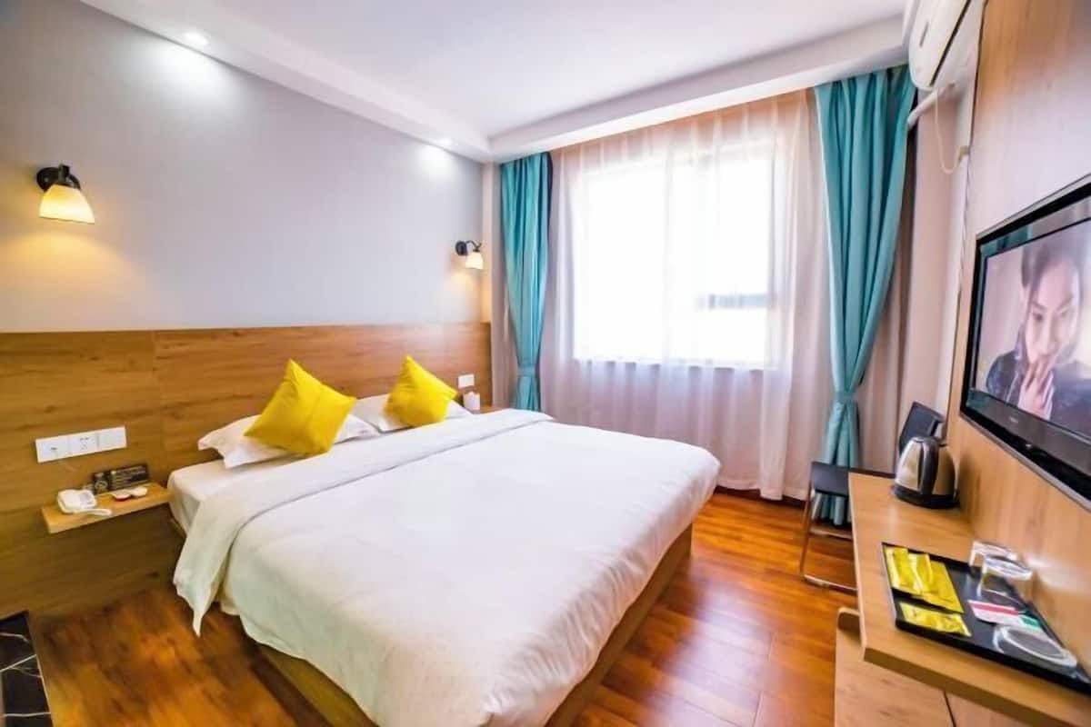 A hotel room with a wooden headboard, a bed with white bedding and two yellow pillows, a bedside table with a phone and a lamp, a TV mounted on the wall, and a window with teal curtains.