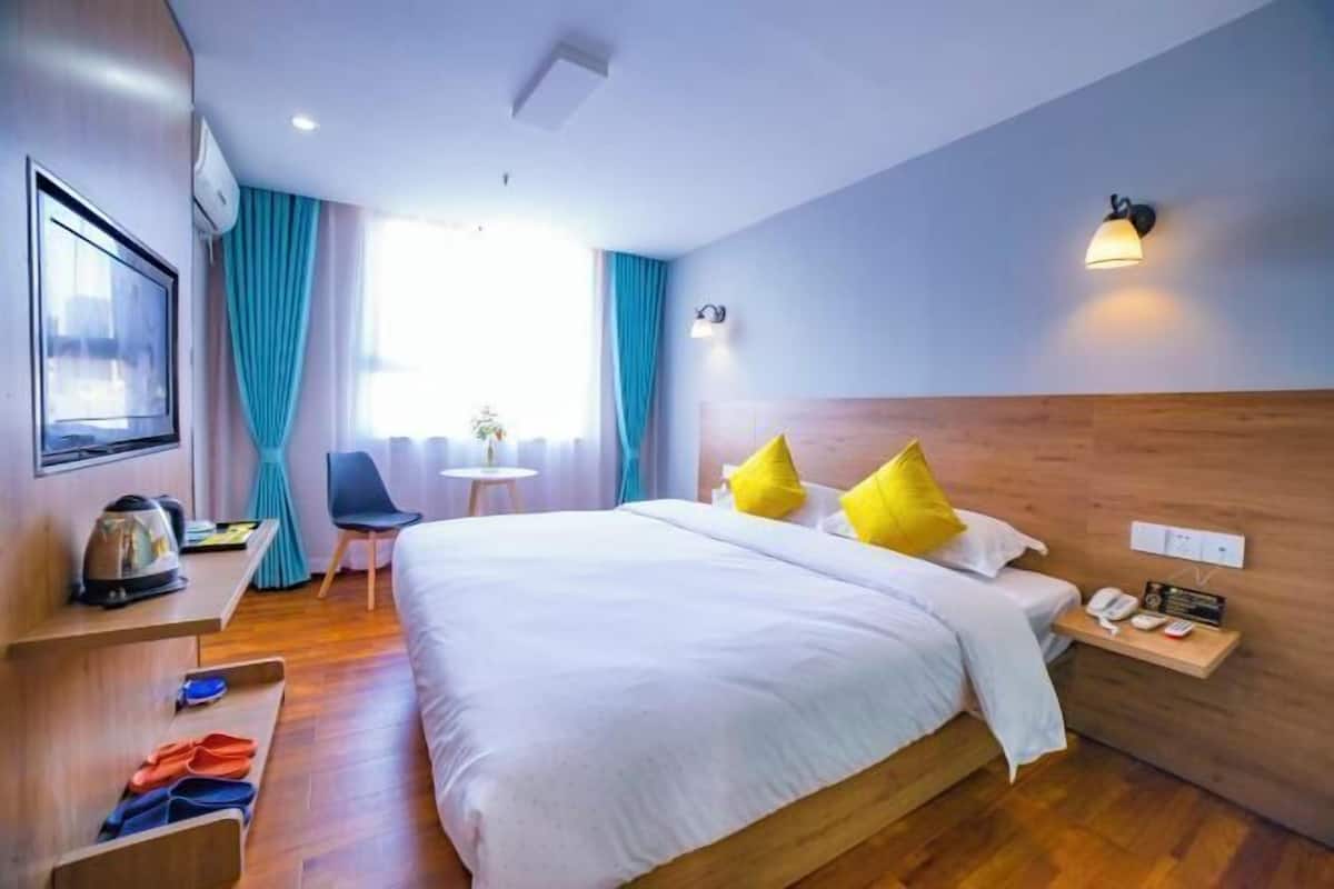 A hotel room with a bed, a desk with a kettle, a chair, a small table, and a wall-mounted phone.
