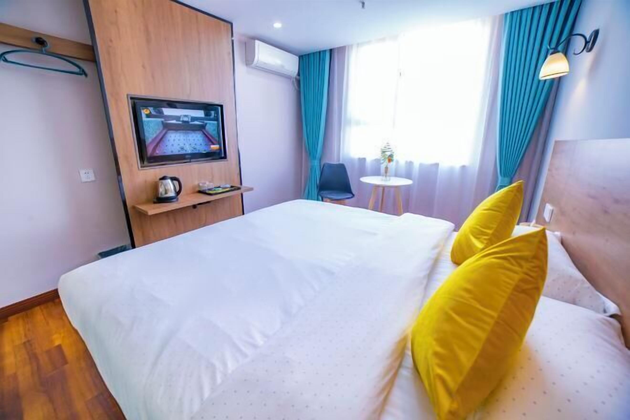 Una habitación de hotel con una cama, un televisor montado en la pared, una mesita con una lámpara y una ventana con cortinas.