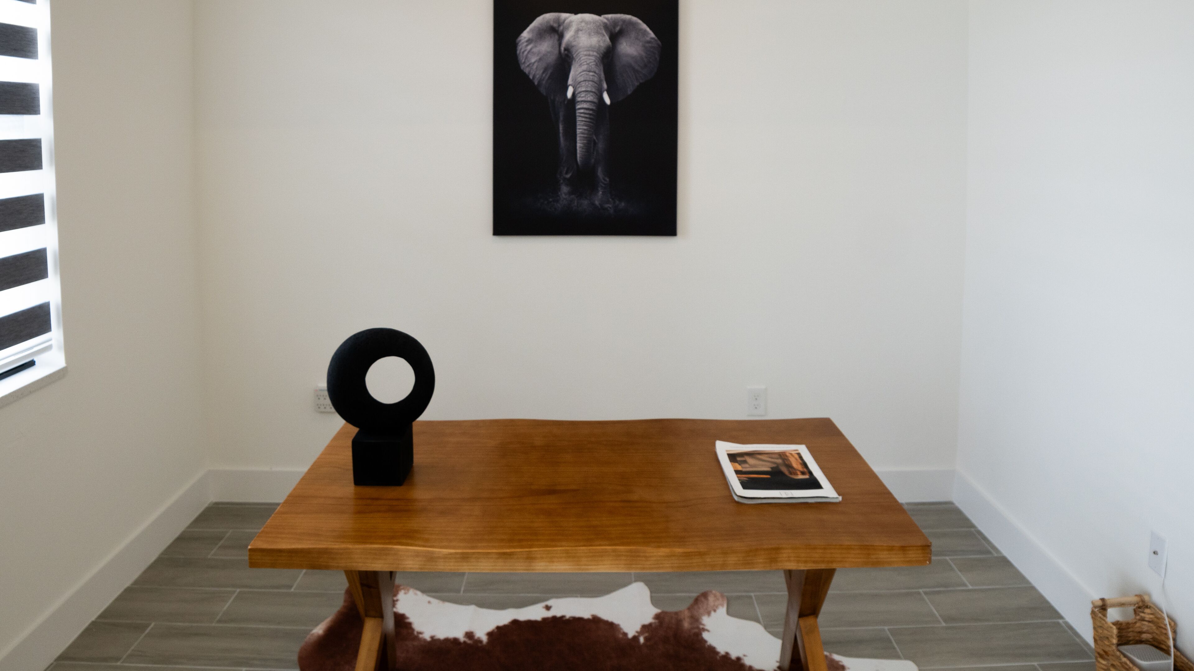 Una habitación con una mesa de madera, un objeto circular negro, un cuadro con un elefante y una alfombra de cuero de vaca.