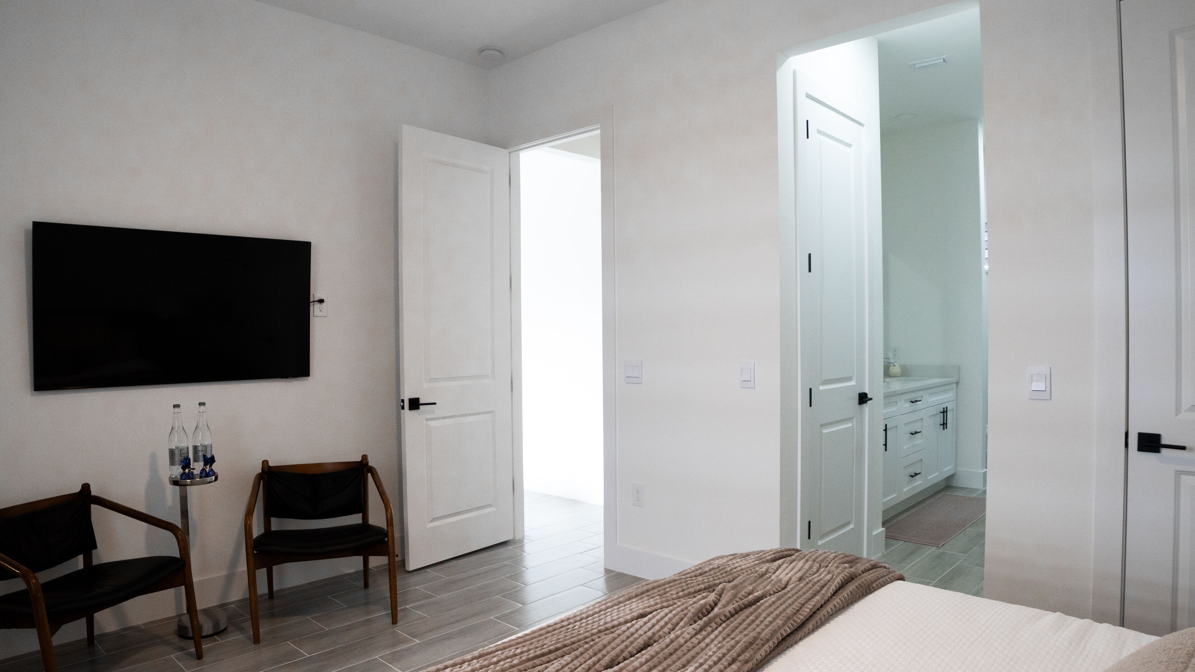A bedroom with a bed, a TV, two chairs, a small table with bottles, and a door leading to a bathroom.