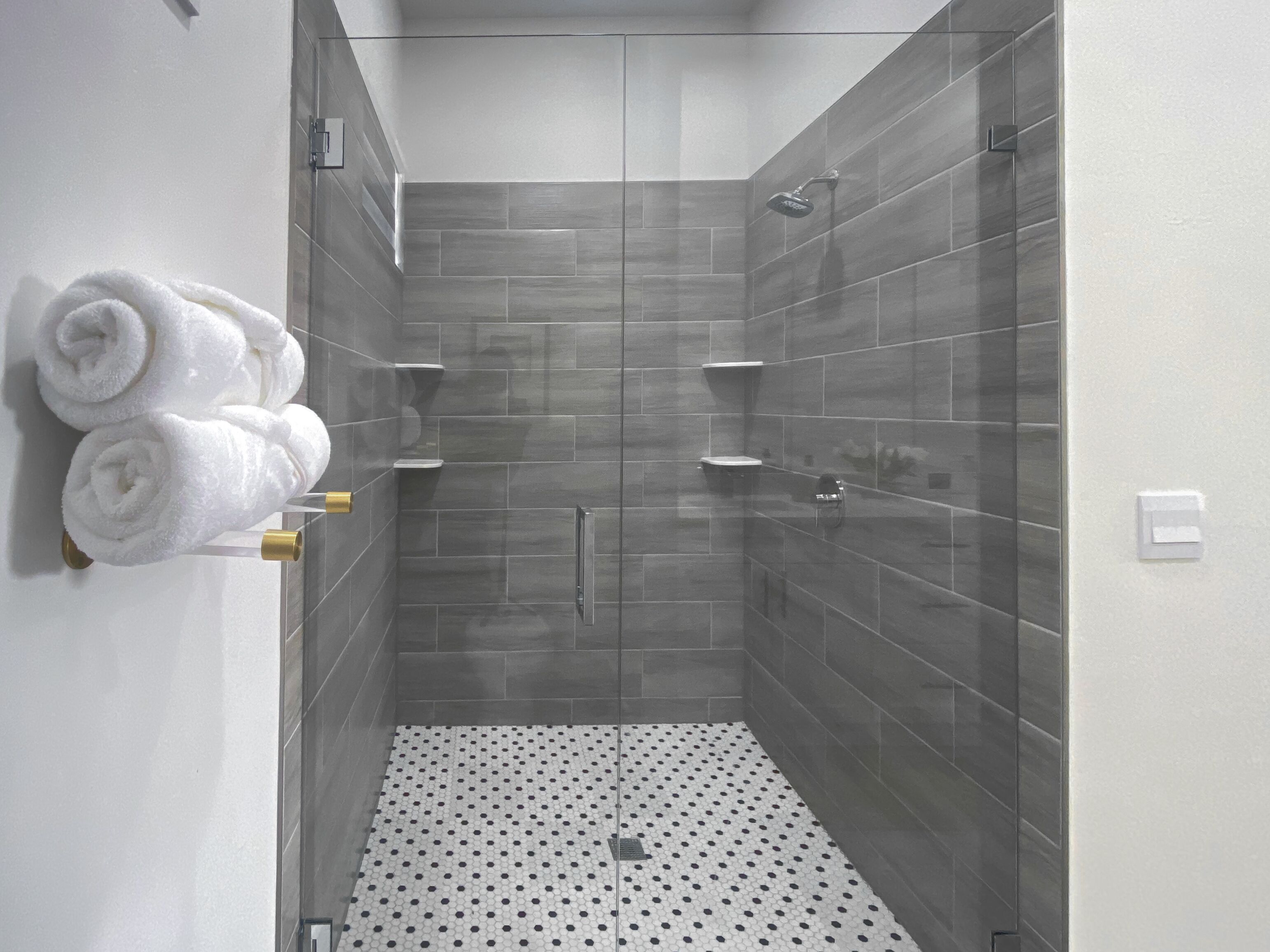 Een moderne douche met grijze tegels, een vloer van witte tegels met zwarte stipjes, en witte handdoeken op een wandrek.