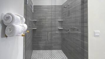 Een moderne douche met grijze tegels, een vloer van witte tegels met zwarte stipjes, en witte handdoeken op een wandrek.