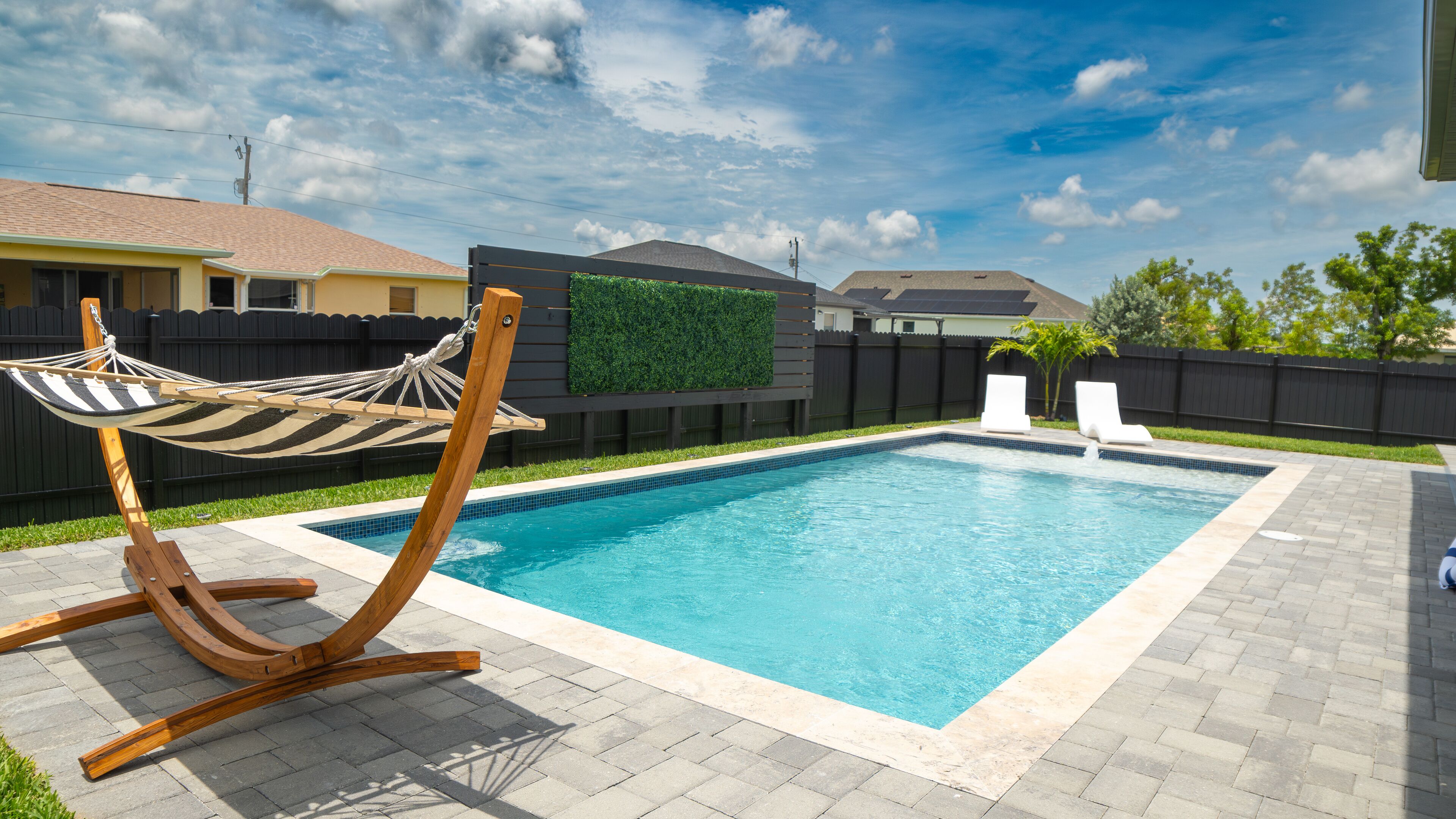 A rectangular swimming pool with a tiled edge, a wooden swing chair, and two white lounge chairs.