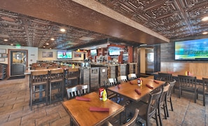 Un restaurant avec des tables et des chaises en bois, un coin bar doté d’un écran de télévision, et un panneau indiquant l’emplacement des toilettes.