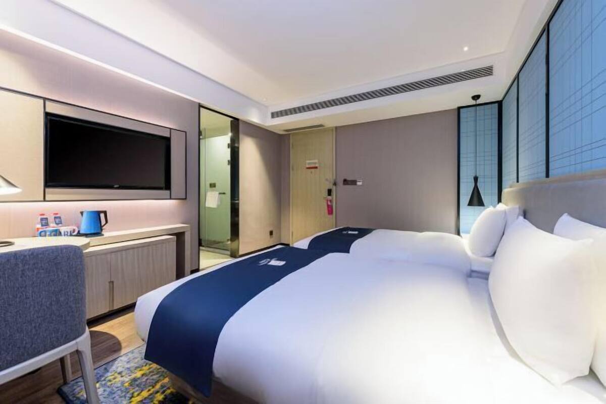 A modern hotel room with a large bed, a desk with a chair, a flat-screen TV, and a large window.
