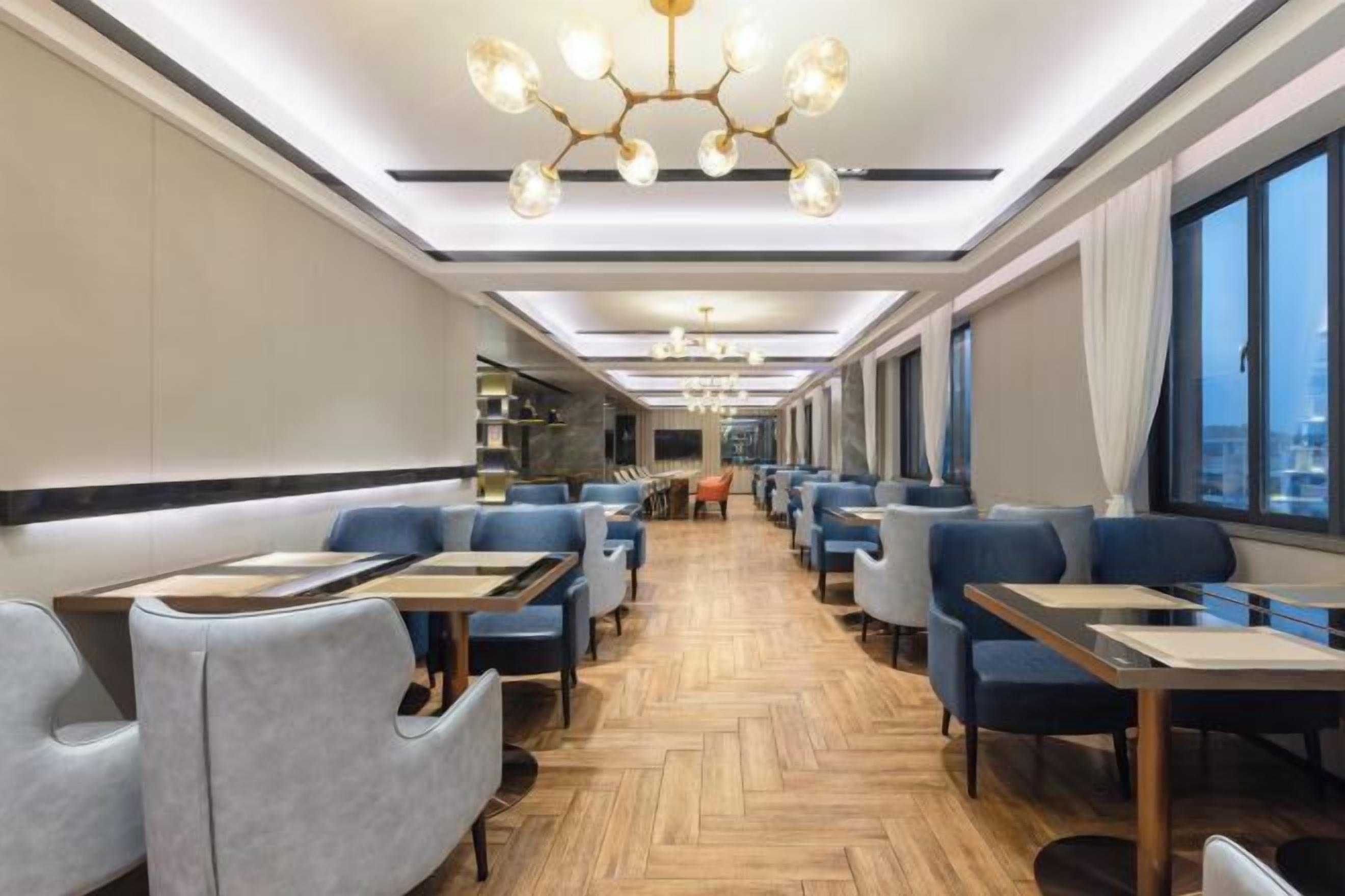 A modern restaurant with a long, central aisle, blue upholstered chairs, and tables with light wood finishes.