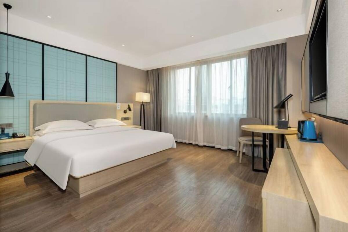 A modern hotel room with a large bed, a desk, a chair, a TV, and a window with curtains.