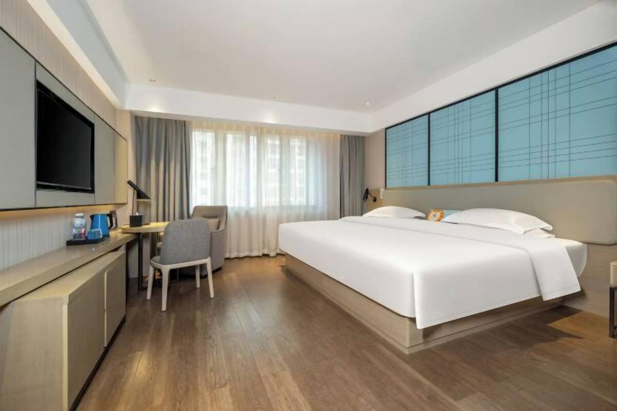 A modern hotel room with a large bed, a desk with a chair, a TV, and a large window with curtains.