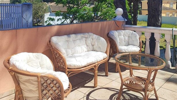 A patio with wicker furniture and a glass table, surrounded by trees and a clear sky.