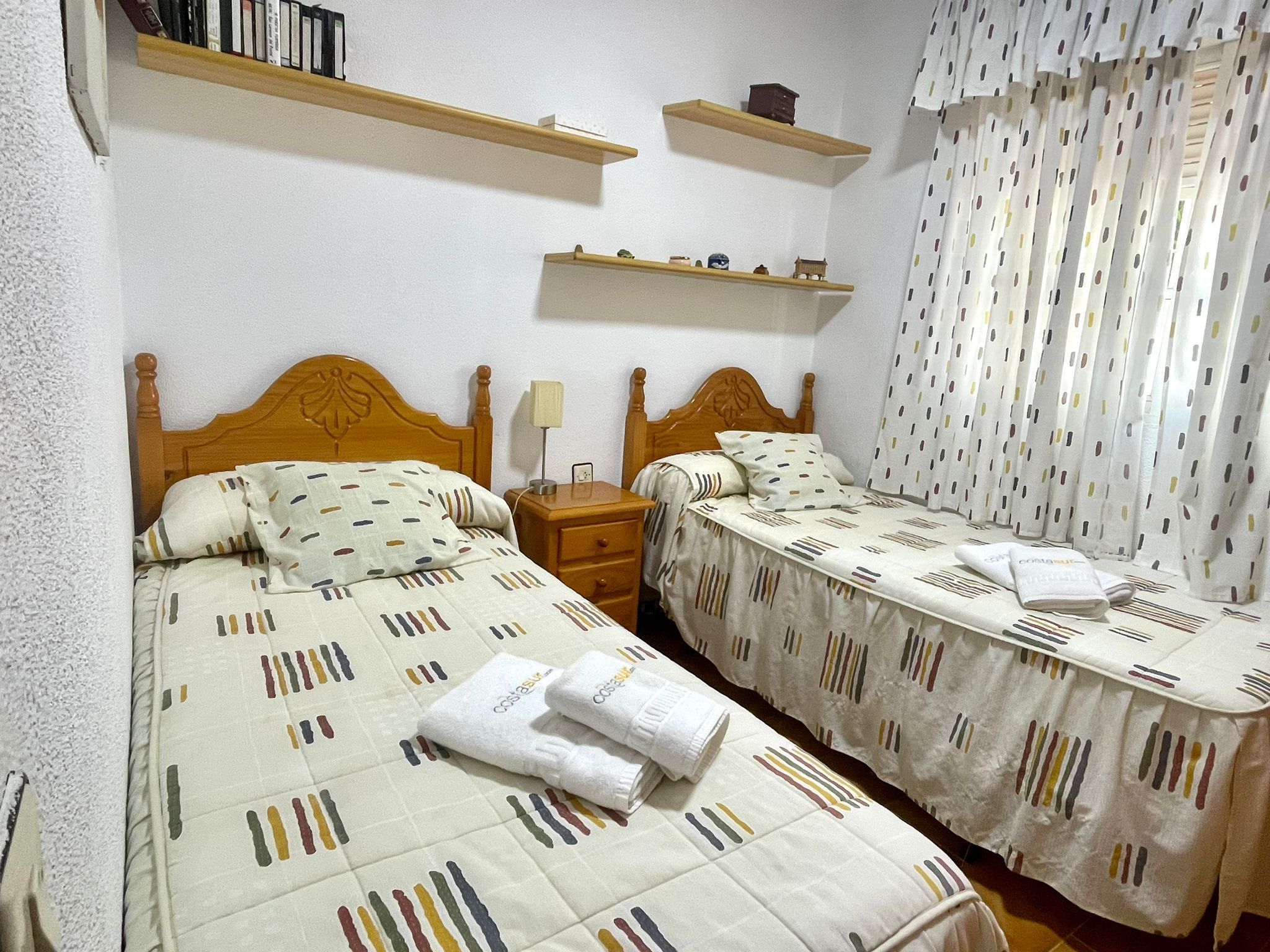 A bedroom with two beds, wooden headboards, bedside tables, and shelves with items.