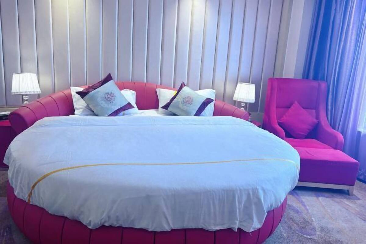 Una cama grande con una colcha blanca y varios almohadones, un sillón tapizado en rojo y una mesita de noche con una lámpara.
