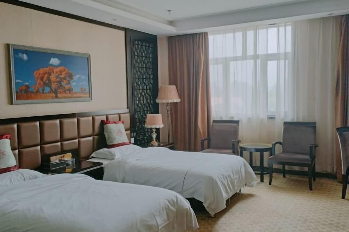 A hotel room with two beds, a small table, a chair, a lamp, and a painting on the wall.