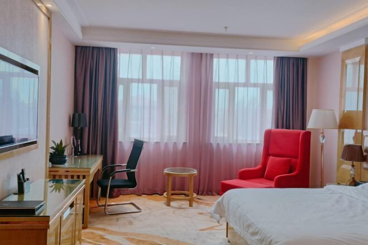 A hotel room with a bed, a desk with a chair, a small round table, and a red armchair.