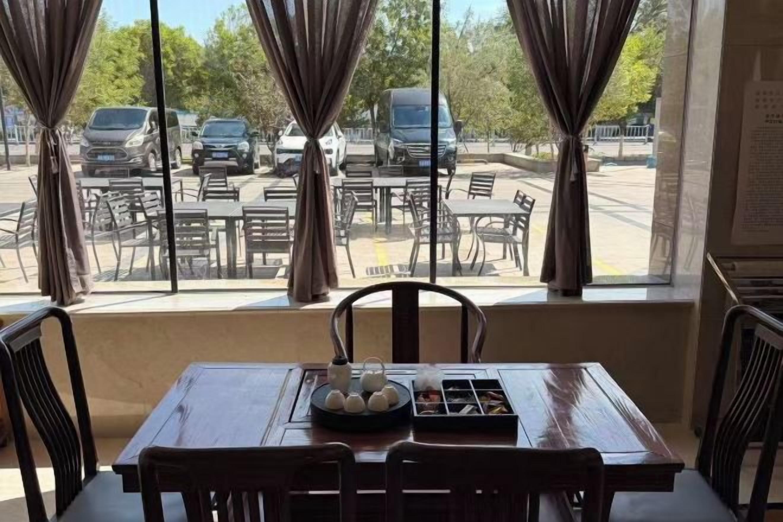 Un coin repas avec une table dressée pour deux personnes, une fenêtre avec des rideaux et une vue sur les voitures garées à l’extérieur.