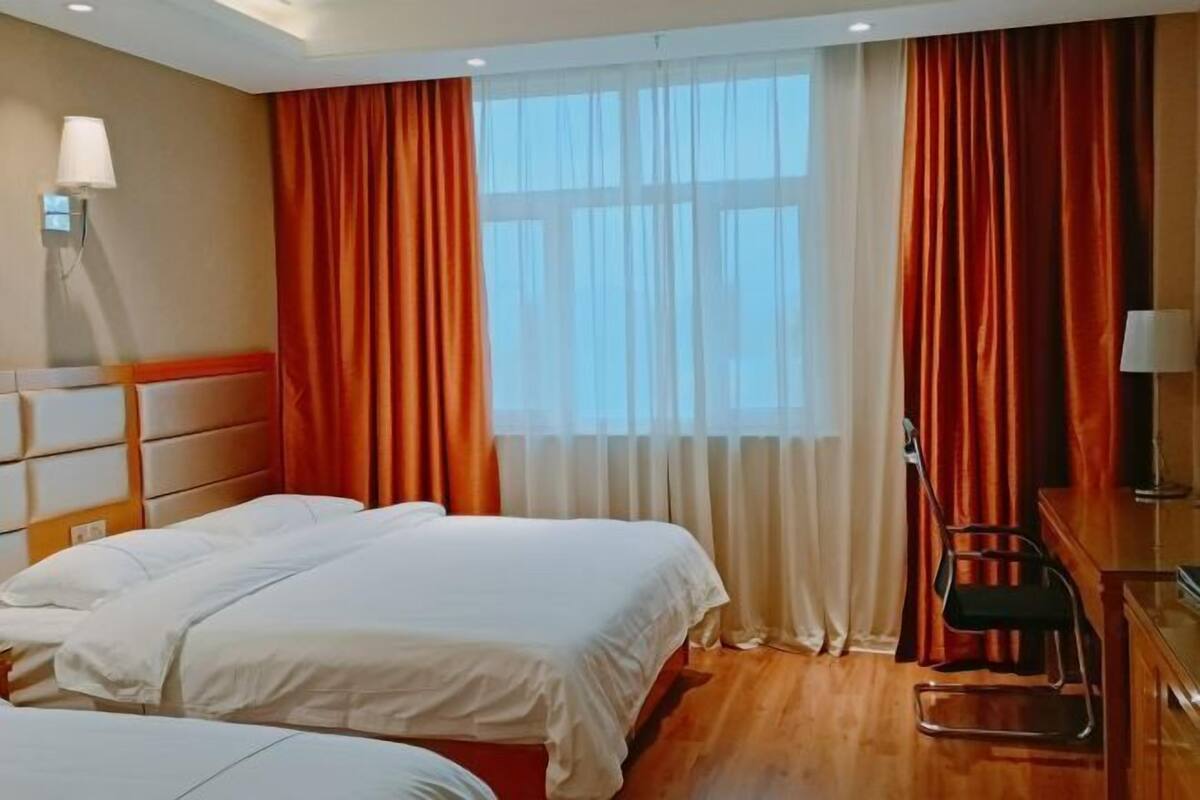 Habitación de hotel con dos camas, un escritorio, una silla y un ventanal grande con cortinas.