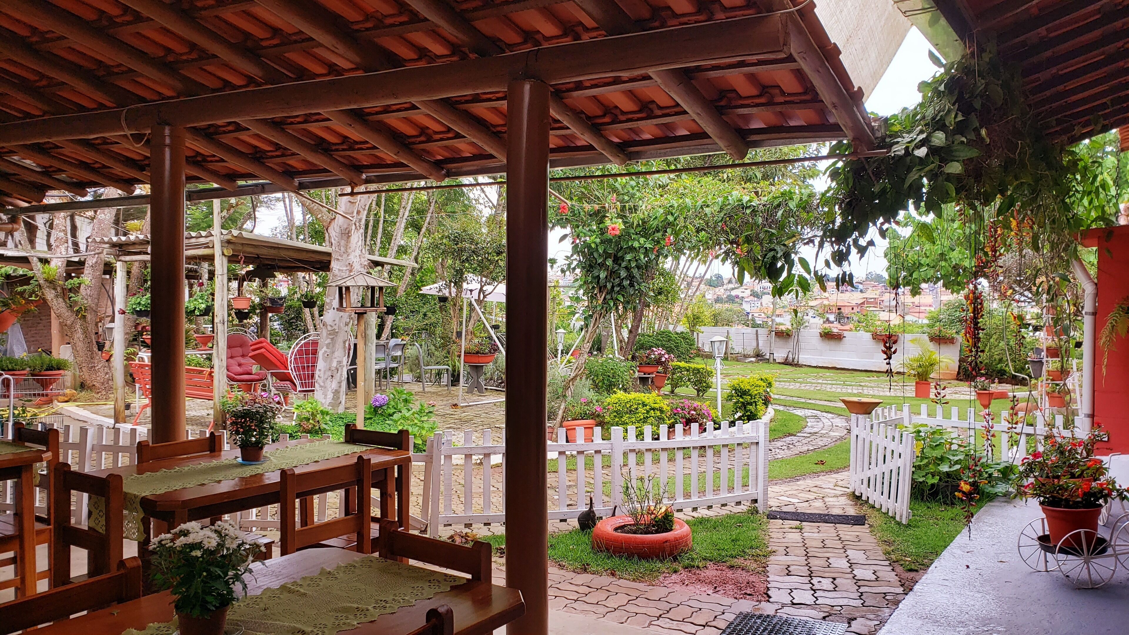 Un espacio de patio cubierto con una mesa y sillas de madera, rodeado de macetas con plantas y una valla de piquetes blanca.