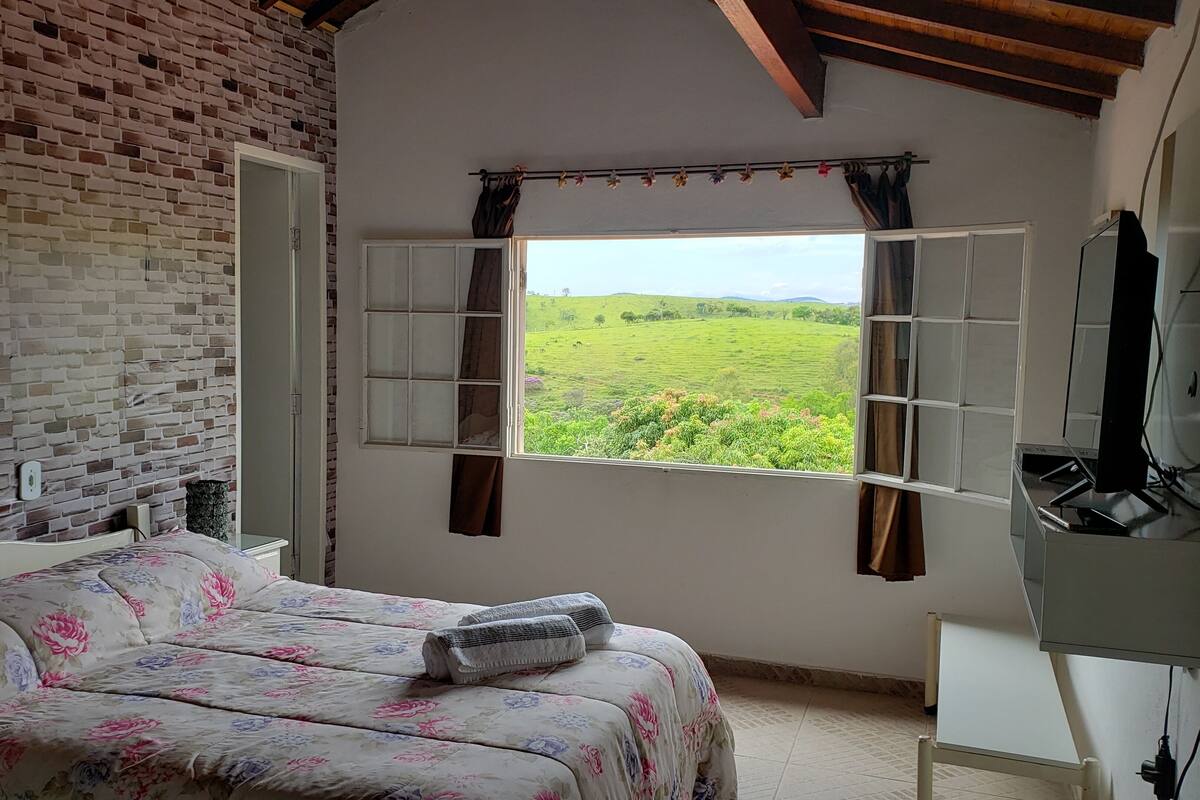 Ein Schlafzimmer mit Backsteinwand, einem Bett mit floralem Bettzeug, einem Fenster mit Vorhängen und einem Fernseher auf einem Schrank.
