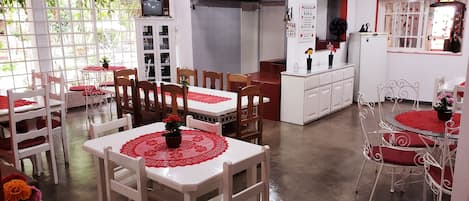 A dining area with white tables and chairs, red tablecloths, and a potted plant on each table.