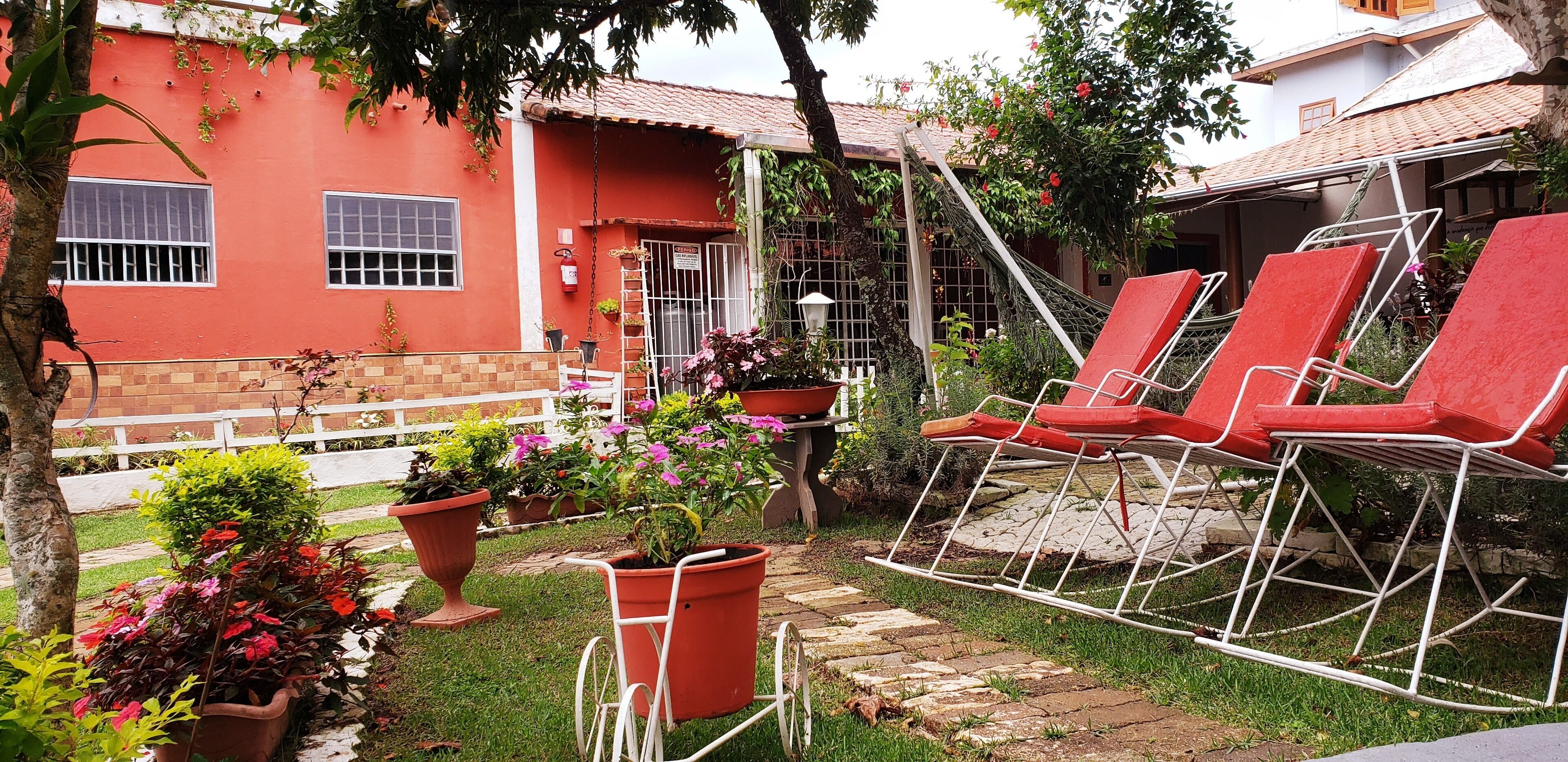 Un edificio rojo con tejado de tejas, cerca blanca y jardín con macetas y sillones rojos.