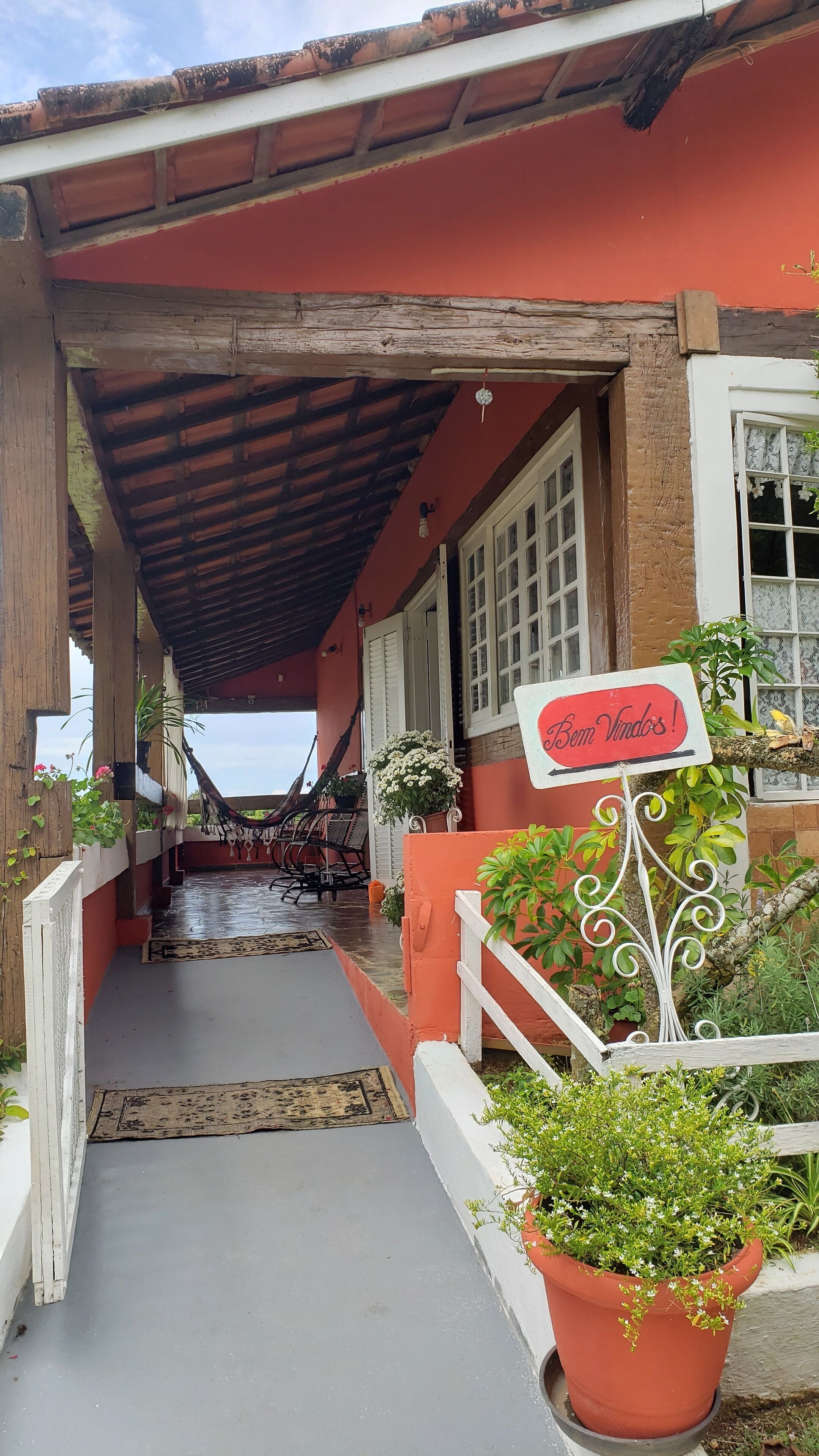 Une entrée couverte menant à un bâtiment à la façade rouge, avec une enseigne indiquant « Bom Vindo! » et des plantes en pot.