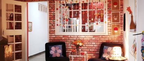A cozy room with a brick wall, two black armchairs, a small table with a vase of flowers, and a large window with decorative elements.
