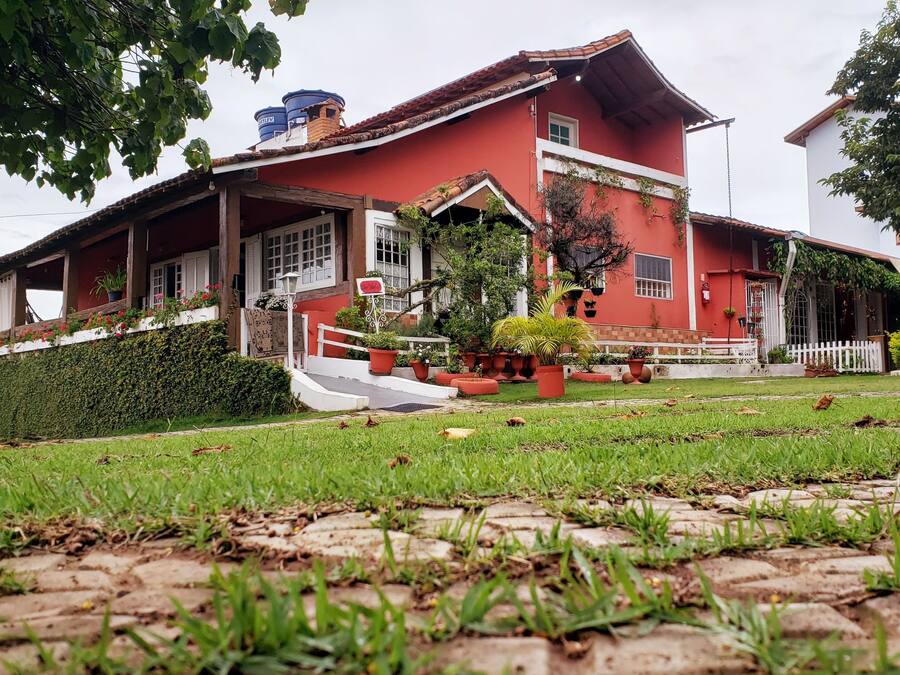 Um edifício vermelho com varanda branca, rodeado por vegetação e vasos de plantas.