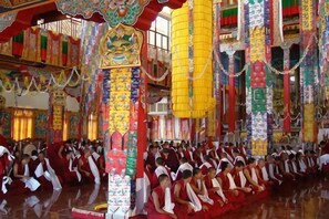 Un amplio salón con filas de personas sentadas, posiblemente monjes, en un entorno tradicional con decoraciones ornamentales.