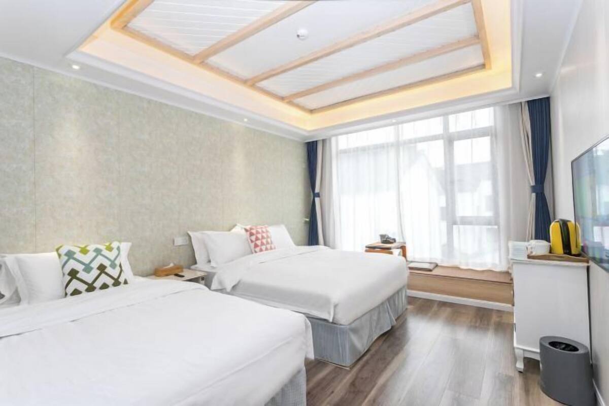 Habitació d'hotel amb dos llits, una finestra gran amb cortines, un sostre amb bigues de fusta i una taula petita amb una làmpada.