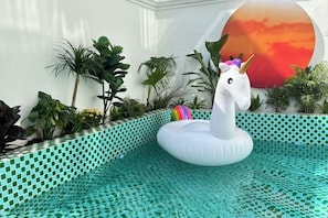 A pool with a white inflatable unicorn float, surrounded by tropical plants and a decorative wall with a sunset mural.