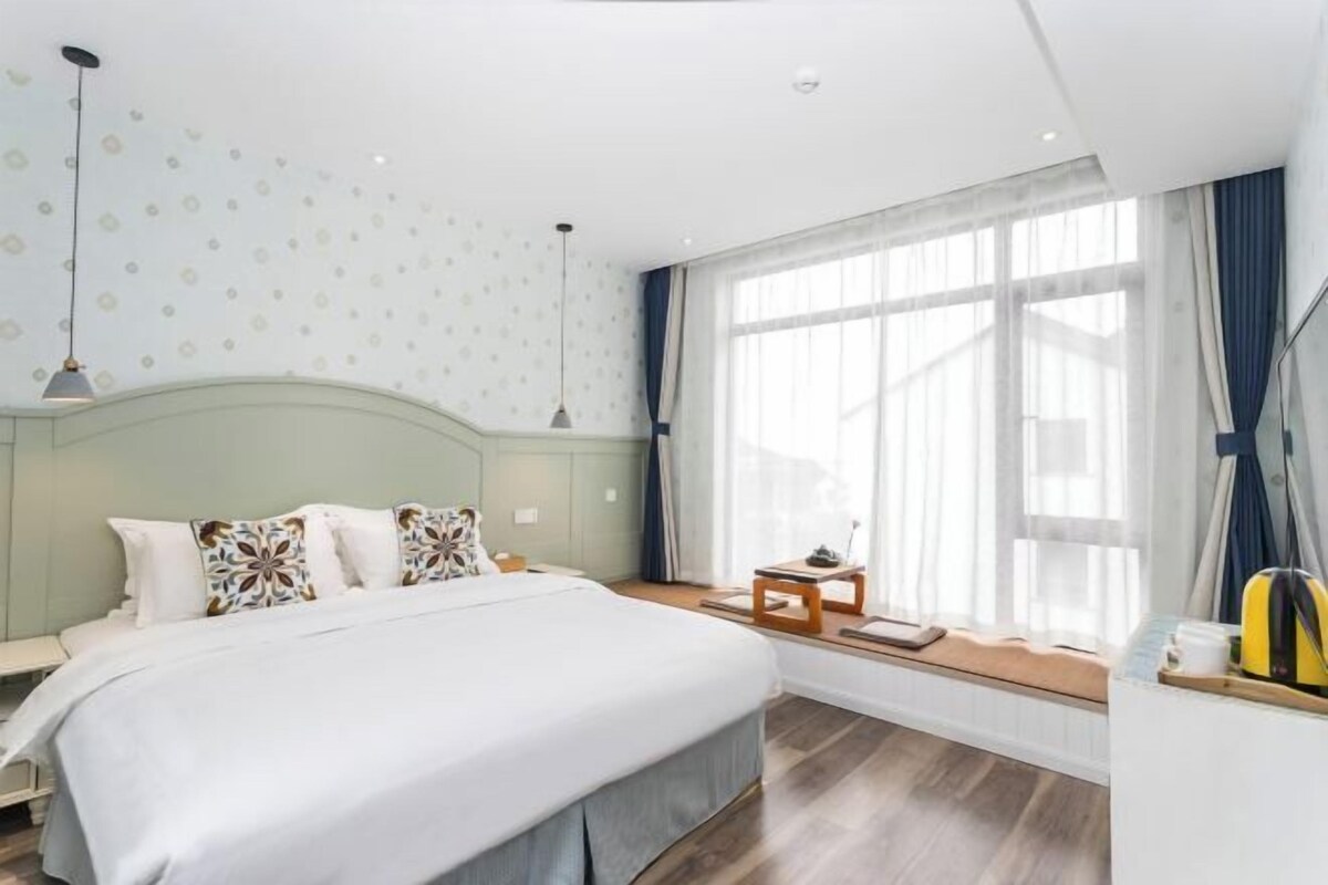 A hotel room with a bed, a small table, a window with curtains, and a ceiling with a light fixture.