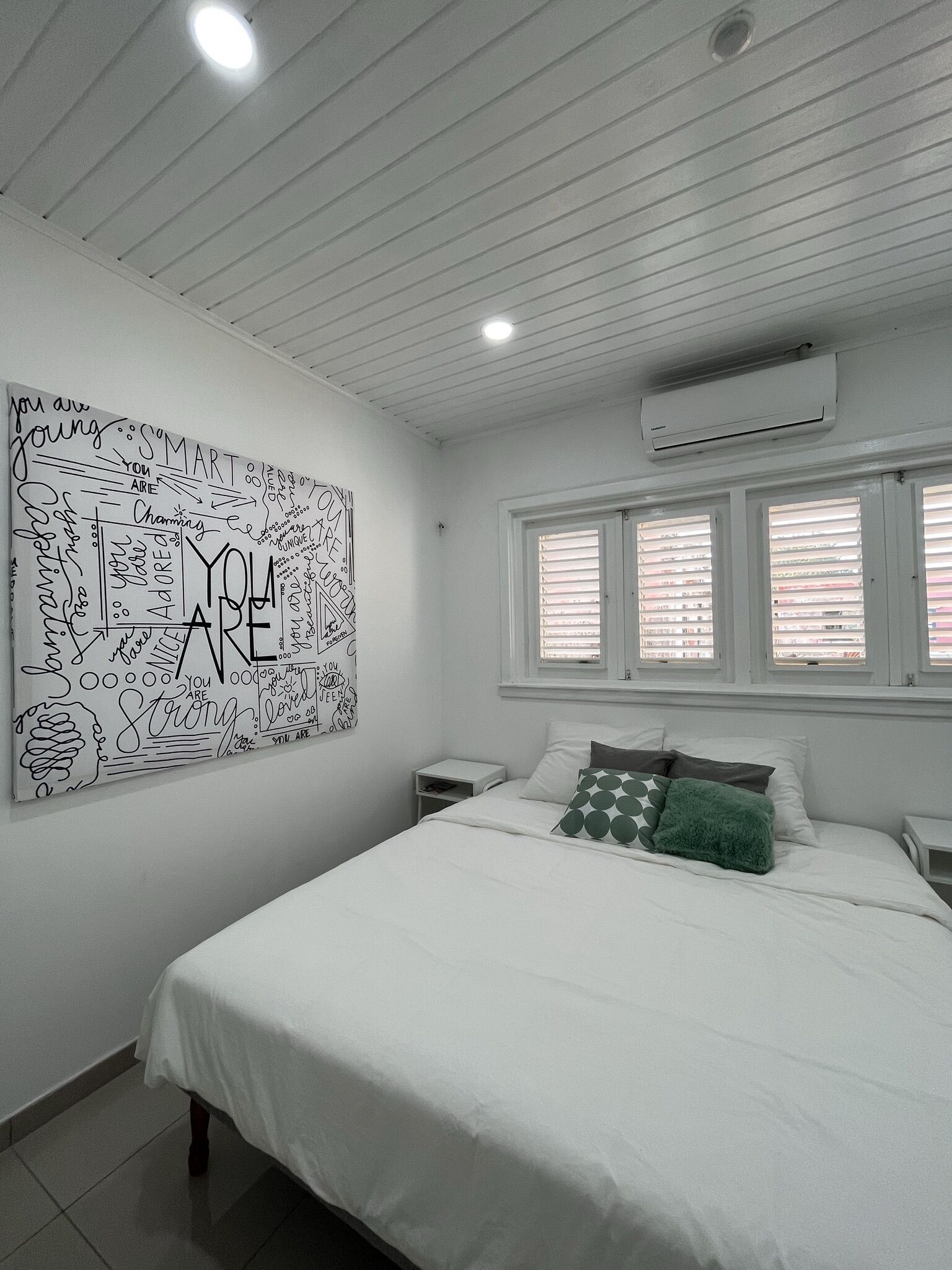 Une chambre avec un grand tableau décoratif aux couleurs vives, un lit avec une literie blanche, une table de chevet et une fenêtre avec des stores.