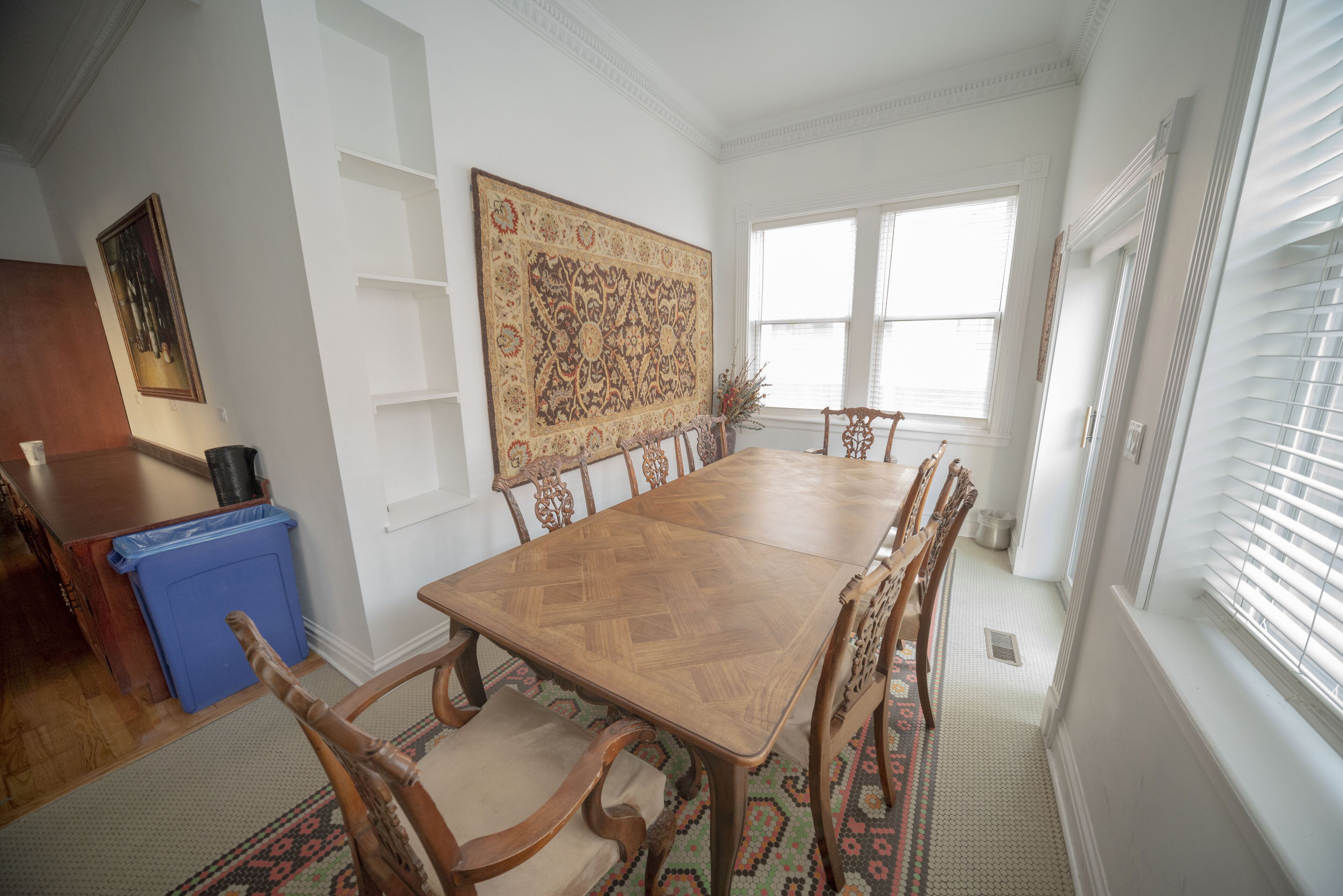 A dining room with a wooden table and chairs, a large patterned rug, and a blue trash bin.