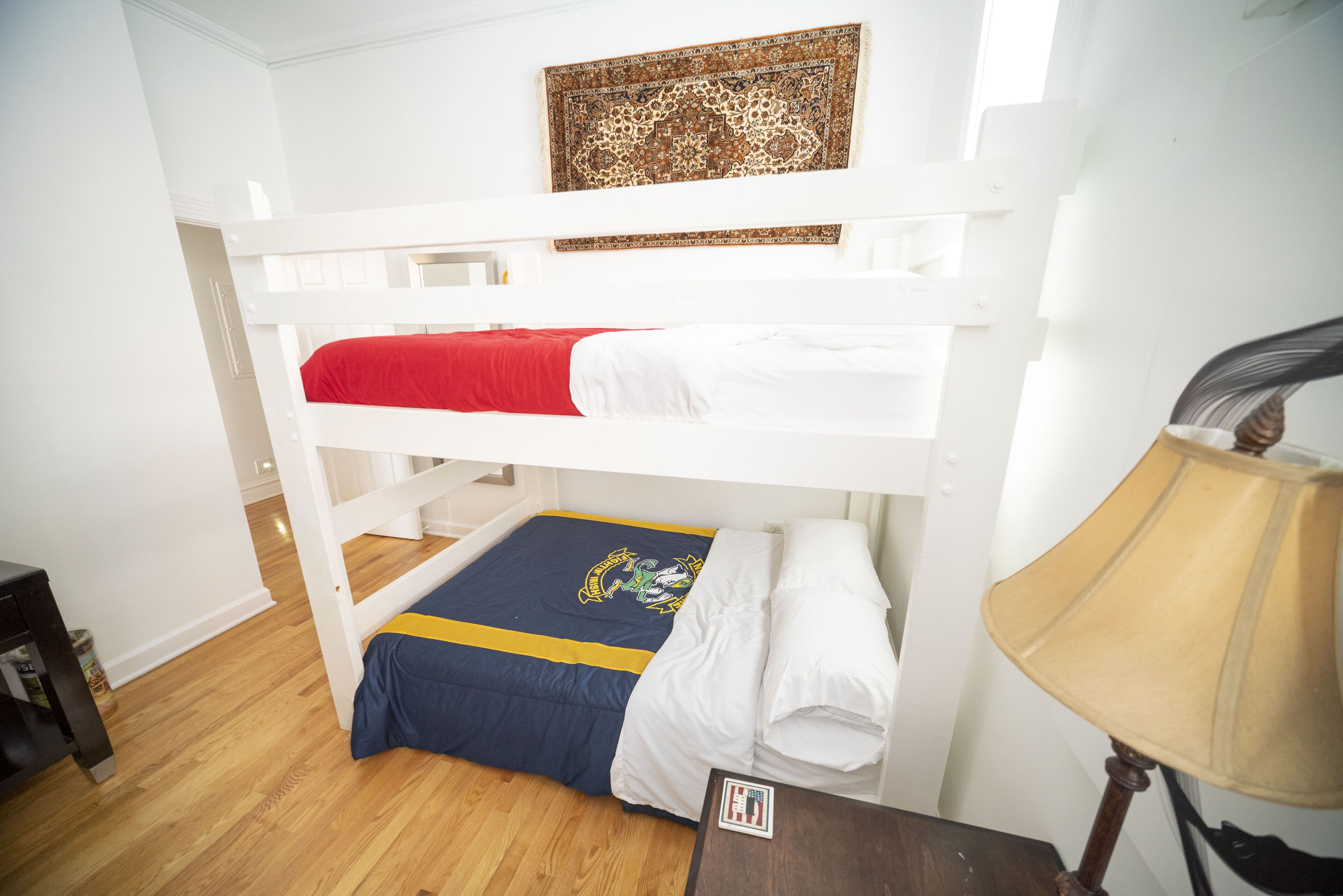 A bunk bed room with a red and blue bedspread, a wooden floor, a lamp, and a decorative wall piece.
