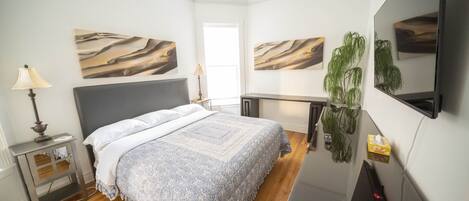 A bedroom with a bed, bedside tables, a desk, a television, and wall art.