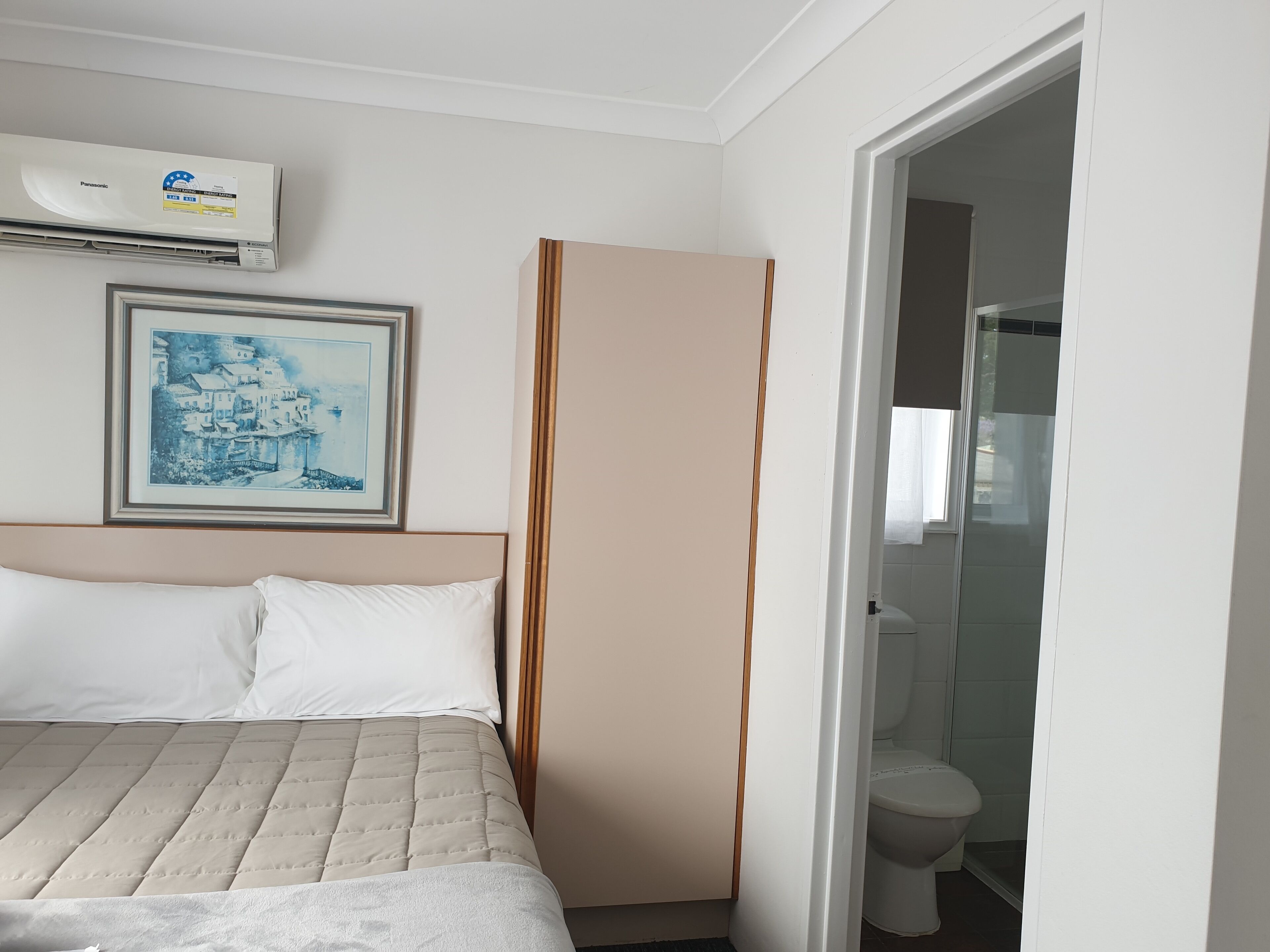 Quarto de hotel com uma cama, um quadro na parede, um ar condicionado e um banheiro visível através de uma porta aberta.
