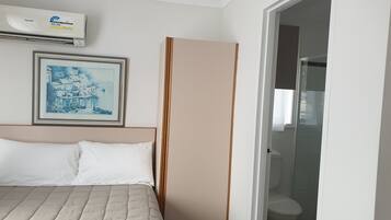 Quarto de hotel com uma cama, um quadro na parede, um ar condicionado e um banheiro visível através de uma porta aberta.
