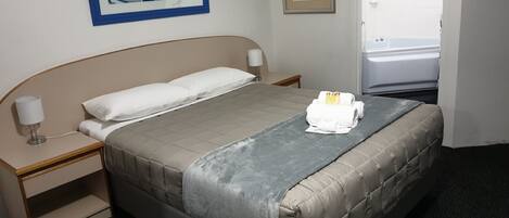 Ein Hotelzimmer mit einem Bett, einem Nachttisch, einer Lampe, einem Bilderrahmen mit einem Strandmotiv und einem Bilderrahmen mit einer Pflanze.