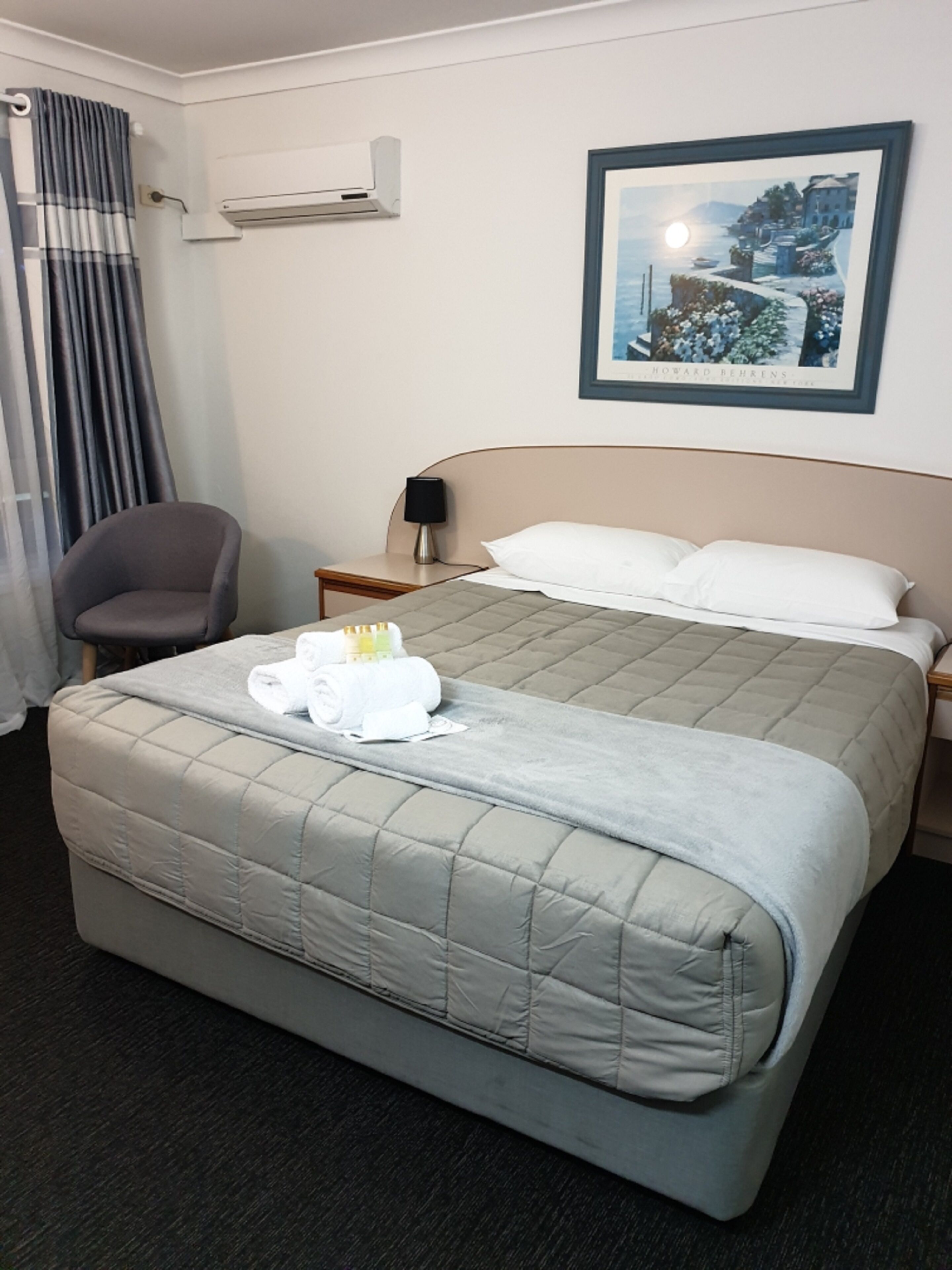 A hotel room with a bed, a chair, a nightstand, an air conditioner, and a framed picture on the wall.