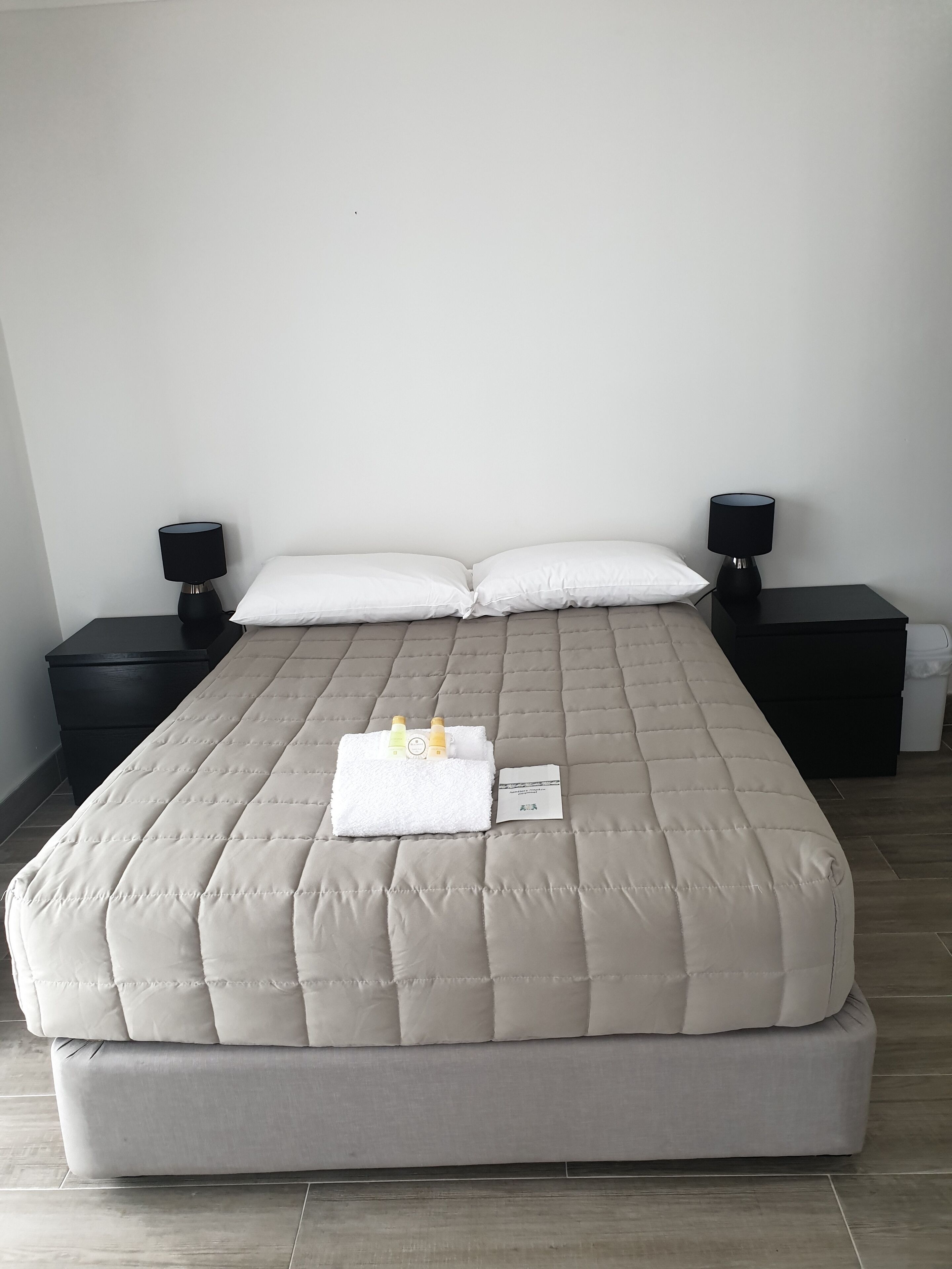 Un lit bien fait, avec une couette blanche, deux oreillers et une serviette pliée, ainsi que des articles de toilette. Il y a deux tables de chevet avec des lampes et une petite poubelle.