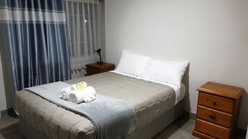 A neatly made bed with a grey bedspread, white pillows, and a wooden nightstand with a lamp.