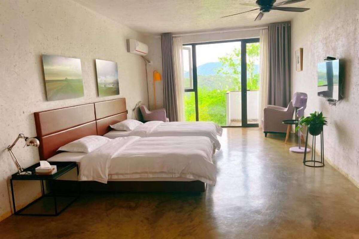 Une chambre d’hôtel avec deux lits, une télévision, un ventilateur de plafond, une grande fenêtre avec vue et un balcon.