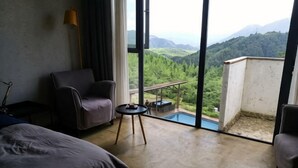 Una habitación con cama, una silla, una mesita y un balcón con vistas a la montaña y la vegetación.