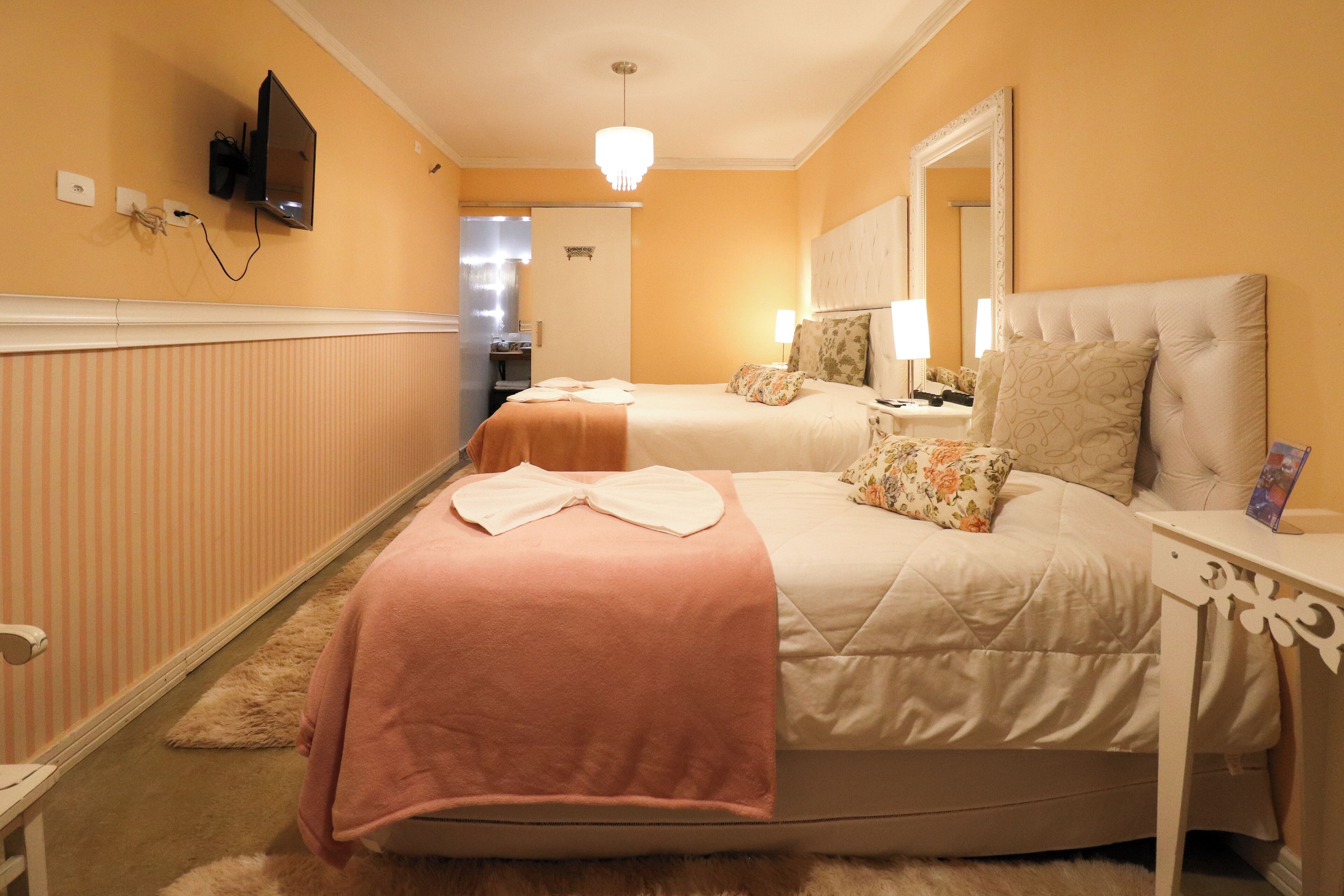 A hotel room with two beds, a television mounted on the wall, a mirror above the bed, and a small table with a lamp.
